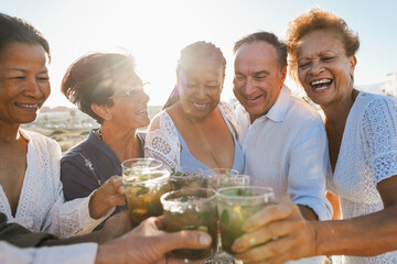 Senior multiracial friends cheering with mojitos on the beach at sunset - Mature people having fun...