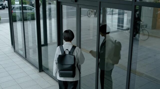 A person in a backpack interacts with a glass door at an entrance, opening it to meet another person inside. The scene is set in a modern, glass-paneled building with outdoor view visible.