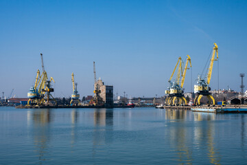 Obraz premium a calm industrial harbor during the day, featuring several yellow portal cranes reflected in the still water under a clear blue sky.