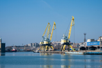 Obraz premium a bustling industrial harbor featuring two prominent yellow portal cranes on a pier, with a city skyline and port buildings in the distance across calm blue water.