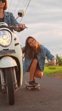 Two young women enjoying an evening ride in Bali, one driving a scooter while the other skateboards along a rural road. Lush rice fields and tall palm trees frame the tropical landscape as the warm