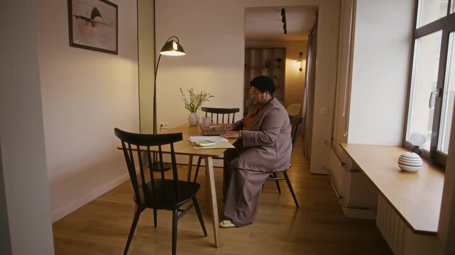 Full slowmo shot of senior African American woman sitting at desk in modern apartment and reviewing financial documents in front of laptop, analyzing monthly expenses