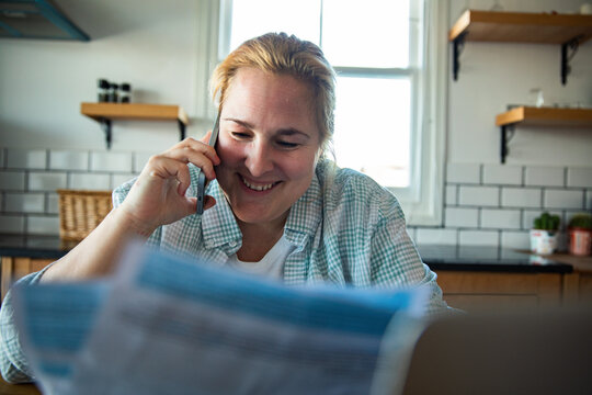 Smiling woman on phone reviewing bills at home kitchen