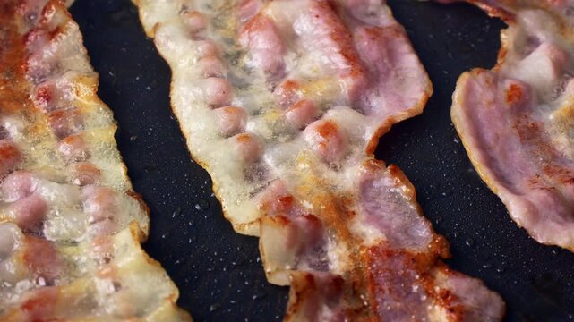 Slices of bacon frying in black pan closeup