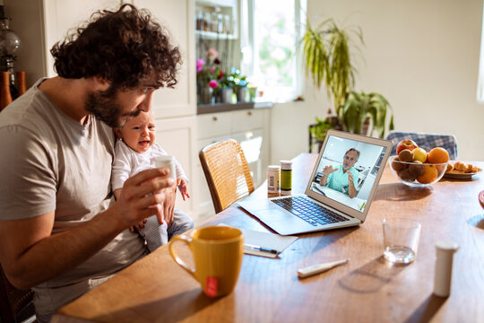Father with baby consulting pediatrician via video call in home kitchen