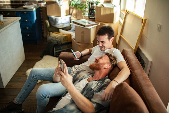 Two men relaxing with smartphones on couch amid moving boxes at home