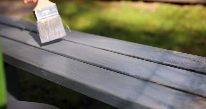man with paintbrush restoring color of garden bench with gray wood protective paint. outdoor wooden furniture protection and renovation