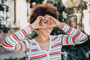 Playful woman frames a heart with hands in a sunny city street smiling bright and stylish today...
