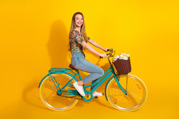 Young woman rides a turquoise bicycle with a wicker basket full of flowers against a bright yellow...
