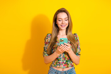 Young woman with a turquoise phone stands against a bright yellow background wearing a floral top...