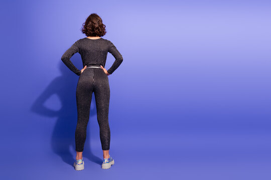Young chic woman in glittery jumpsuit stands with hands on hips against purple studio background for fashion lifestyle and fitness shoot