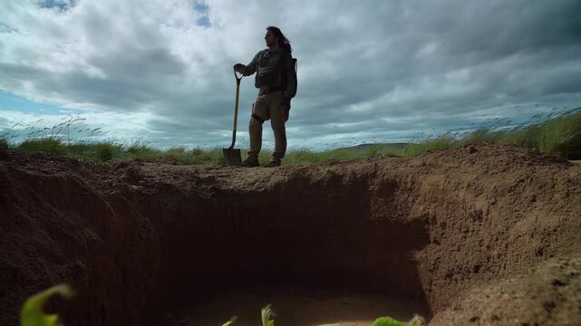Man standing on edge of deep hole with shovel in hand under cloudy sky.