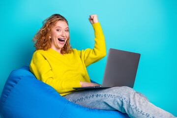Young red hair woman with laptop sits on blue bean bag celebrating success and happiness in a...