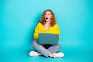 Young woman in yellow sweater with laptop on bright blue background looks surprised and inspired