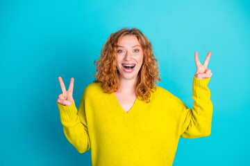 Young redhead woman in a yellow sweater smiles and shows peace signs against blue background