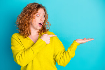 Young redhead woman in yellow sweater points to open palm with surprised expression for stock photo