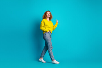 Young woman in bright yellow sweater walks against turquoise background smiles and shows peace sign