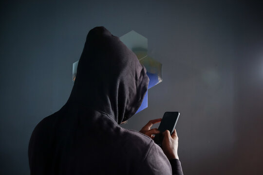 Anonymous man wearing black hoodie using smartphone indoors with his back to the camera, Phone Scammers, mysterious person typing message on mobile phone