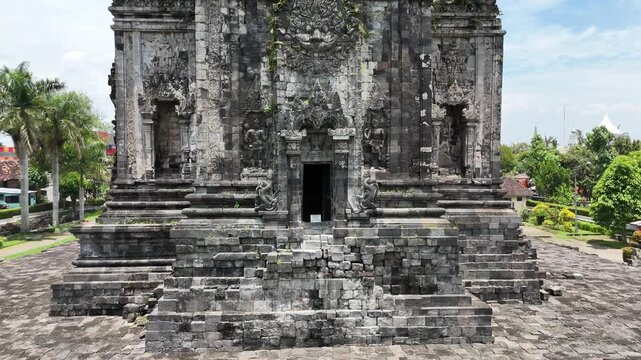 Aerial view of Candi Kalasan Buddhist temple in Yogyakarta, Indonesia. Drone flying upwards along the stone facade of the ancient structure during sunny day.

