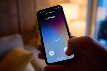 Close-up of hand holding smartphone showing unknown incoming call in living room. Concept of spam...