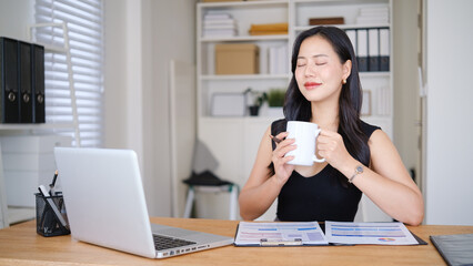 Businesswoman enjoying a coffee break at her desk, holding a mug with eyes closed while working...