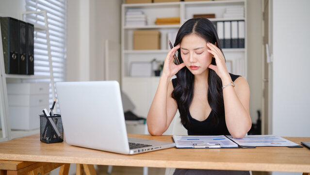 Stressed businesswoman reviewing financial reports at her desk, touching her temples while working on a laptop in a modern office.