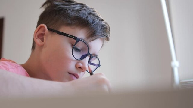 Young boy with glasses concentrating on homework, pencil in hand, focused on writing or drawing at his desk during study time at home, thoughtful and studious