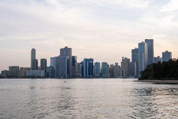 Obraz premium 12.12.2025 Sharjah, UAE. Panoramic view of Sharjah Corniche at sunset, featuring Khalid Lagoon water, illuminated skyscrapers, modern city skyline, and silhouette of date palm trees in UAE
