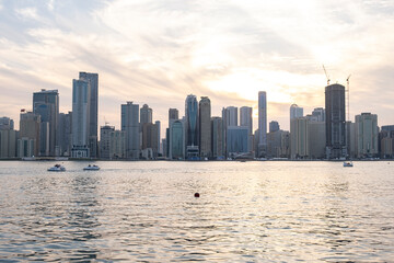 Obraz premium 12.12.2025 Sharjah, UAE. Panoramic view of Sharjah Corniche at sunset, featuring Khalid Lagoon water, illuminated skyscrapers, modern city skyline, and silhouette of date palm trees in UAE