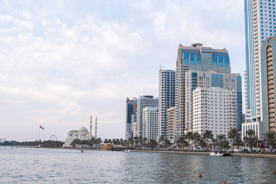 12.12.2025 Sharjah, UAE. Panoramic view of Sharjah Corniche at sunset, featuring Khalid Lagoon water, illuminated skyscrapers, modern city skyline, and silhouette of date palm trees in UAE