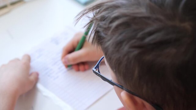 Young boy with glasses seated at a desk, concentrating as he writes homework in a ruled notebook with a green pen, focused on learning and study tasks at home