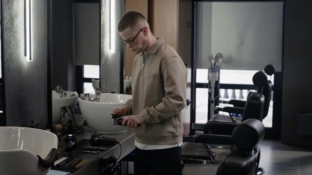 Young Caucasian barber using brush to clean his electric razor while preparing for next client in spacious salon