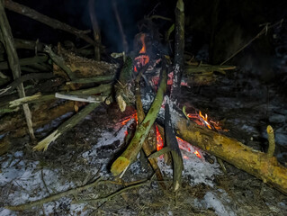 Ognisko na zimowym kampingu w lesie. Ognisko otoczone nieregularnie ułożonymi kłodami drewna w zimowej scenerii lasu. Ogień płonie pomarańczowymi i czerwonymi żarami, rzucając ciepłą poświatę . © Grzegorz