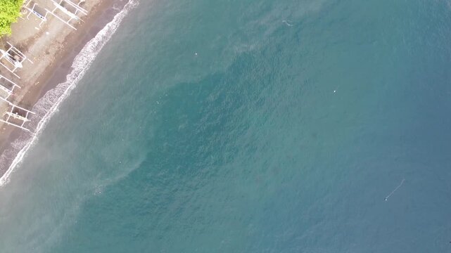 Top-down aerial drone tilting up from ocean water to reveal Amed coastline and Mount Agung volcano in Bali, Indonesia.