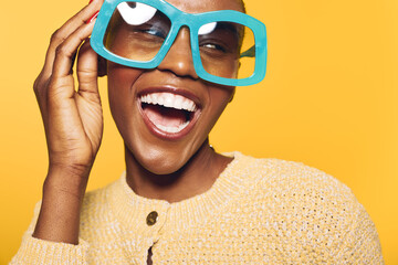 Happy dark skinned African woman wearing oversized blue glasses laughs joyfully on yellow...