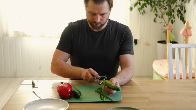 Man preparing fresh vegetables in modern kitchen, organic ingredients. Healthy meal. Cooking Food At Home. Male slicing red pepper and green cucumber on a wooden cutting board. Nutritionist, culinary