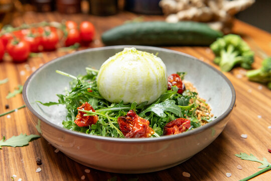 Gourmet burrata salad served in a rustic gray bowl. The creamy burrata sits on a bed of fresh arugula, topped with a drizzle of olive oil.