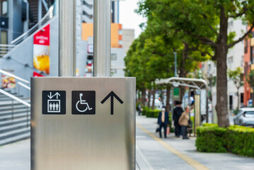 A stainless steel signage with elevator and wheelchair accessibility symbols on a city sidewalk. Concept of inclusive urban design, wayfinding, and modern public infrastructure in Japan.