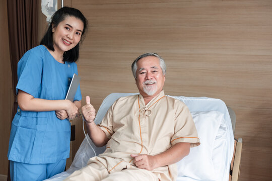 Asian nurse standing beside senior hospital patient giving thumbs up while sitting on hospital bed, healthcare support and positive recovery concept in medical environment