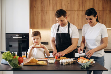 Family cooking together in a modern kitchen, a man and woman prepare vegetables while a boy enjoys a snack at the kitchen island with fresh ingredients around © standret