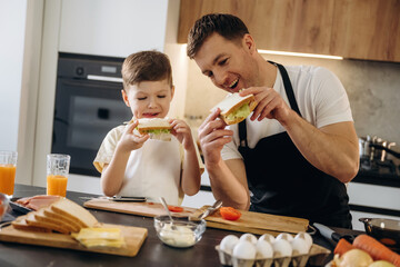 Father and son preparing sandwiches together in a modern kitchen with fresh ingredients, including bread, vegetables, and drinks on the countertop