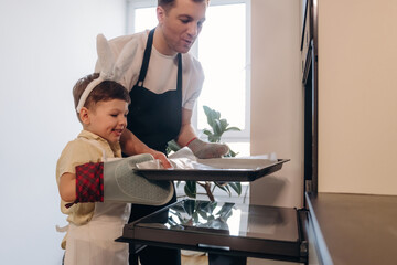 Adult male and young boy wearing aprons and oven mitts take a baking tray out of an oven in a modern kitchen with plants and natural light