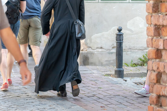 Unidentified catholic clergy member in black cassock walking on city street, back view with shoulder bag, people around on sidewalk, religion christianity priest concept, copy space, no face
