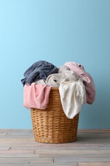 Wicker laundry basket overflowing pastel garments against blue wall on wooden floor, casual household scene