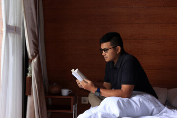 Young Asian male Wearing Glass Reading A Book on the Bed