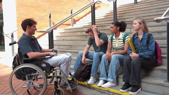 Group of diverse college students relaxing and talking together outdoors. A young man in a wheelchair is included in the friendly conversation