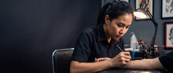 Asian female tattoo artist drawing on a client's arm in a studio. Professional woman at work in a tattoo parlor. Creative occupation. Copy space