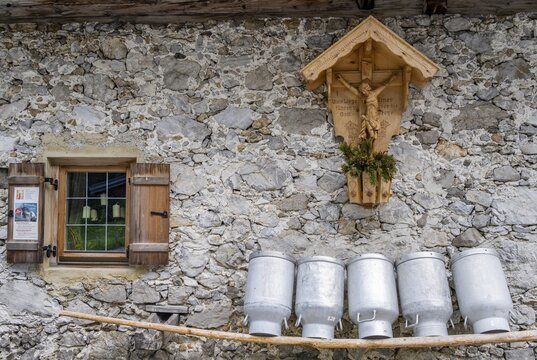 Milk cans lined up on a wooden bench, window and shrine with Jesus on the cross, fa&ccedil;ade made of rough stones from an alpine pasture, Gramaialm, Alpenpark Karwendel, Tyrol, Austria