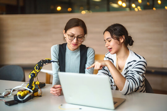 Two young women collaborating on a robotics project with a laptop and robotic arm