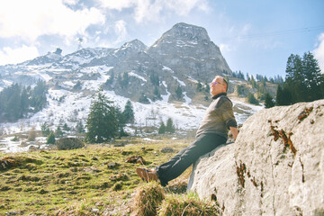 Naklejka premium Senior man relaxing on rock in Swiss Alps near Gruyere, enjoying sunny mountain landscape with snow peaks, peaceful travel lifestyle and nature freedom